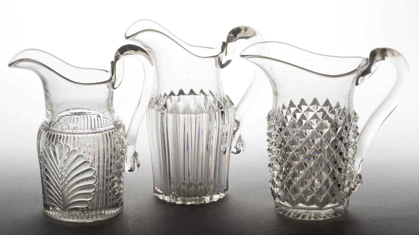 ASSORTED FLINT EAPG WATER PITCHERS, LOT OF THREE: ASSORTED FLINT EAPG WATER PITCHERS, LOT OF THREE, colorless lead glass, comprising a Sawtooth, a Triangular Prism, and a Fern, each having an applied handle and polished pontil mark. Various makers.&n