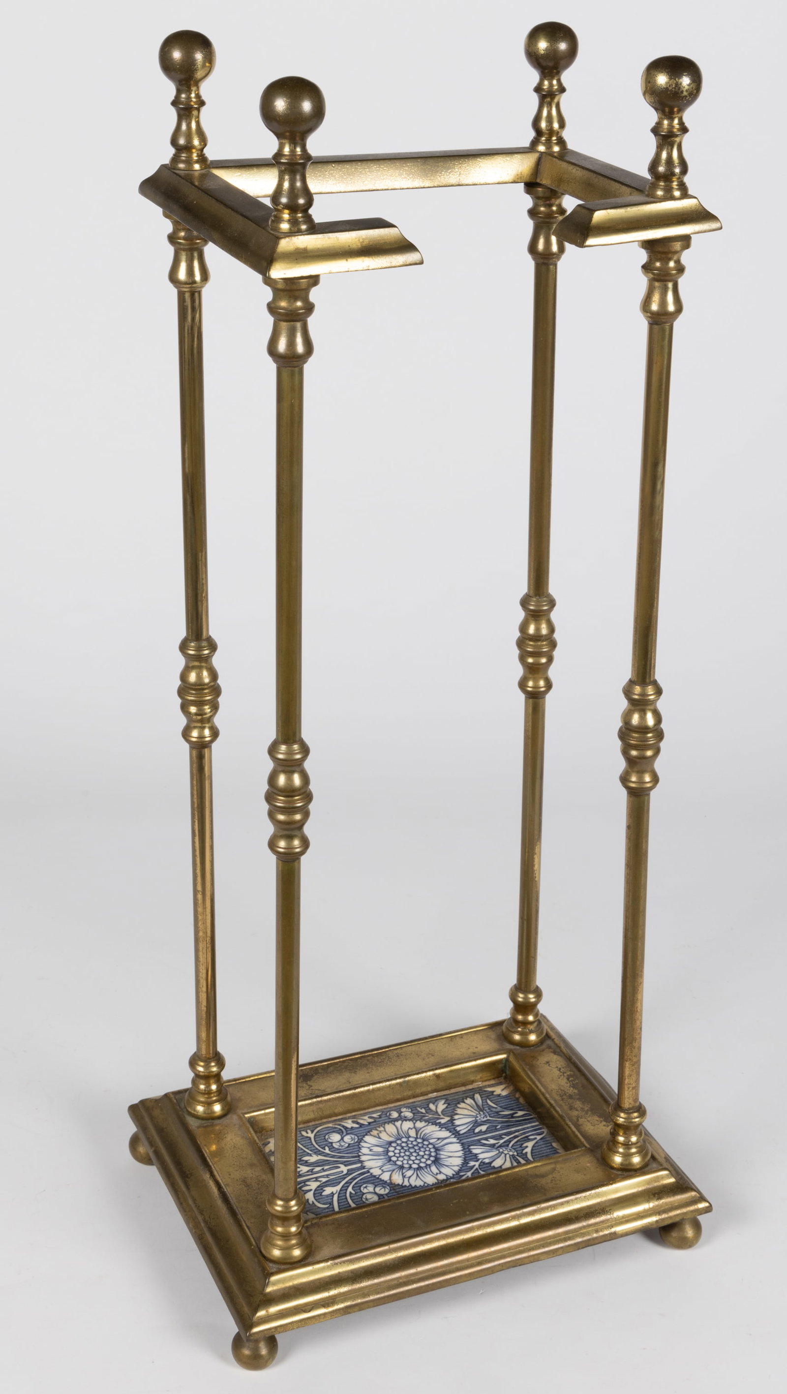 ANTIQUE BRASS AND CERAMIC UMBRELLA STAND: ANTIQUE BRASS AND CERAMIC UMBRELLA STAND, C-form top supported on four posts resting on a bottom frame set with aa Aesthetic Movement blue and white transfer ceramic tile featuring a stylized sun