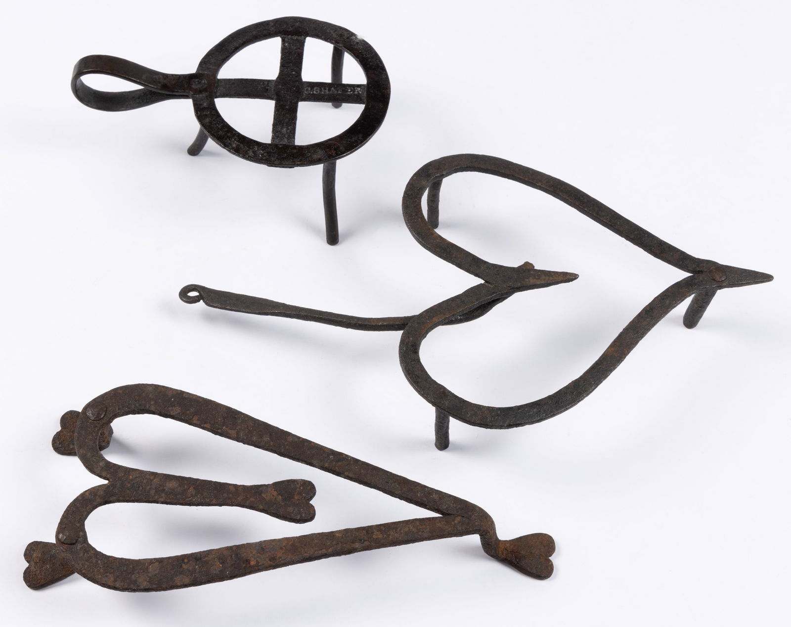 WROUGHT-IRON FIGURAL HEART HEARTH TRIVETS, LOT OF TWO: WROUGHT-IRON FIGURAL HEART HEARTH TRIVETS, LOT OF TWO, comprising one with rotating handle, and one raised on three legs terminating in heart-shaped feet, and interior terminating in a heart, no