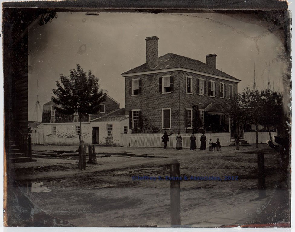 AMERICAN SEAPORT FULL-PLATE AMBROTYPE: AMERICAN SEAPORT FULL-PLATE AMBROTYPE, depicting a brick Georgian home with two tall end chimneys, wooden shutters and slate roof, six African American figures standing at the white picket fence, resi