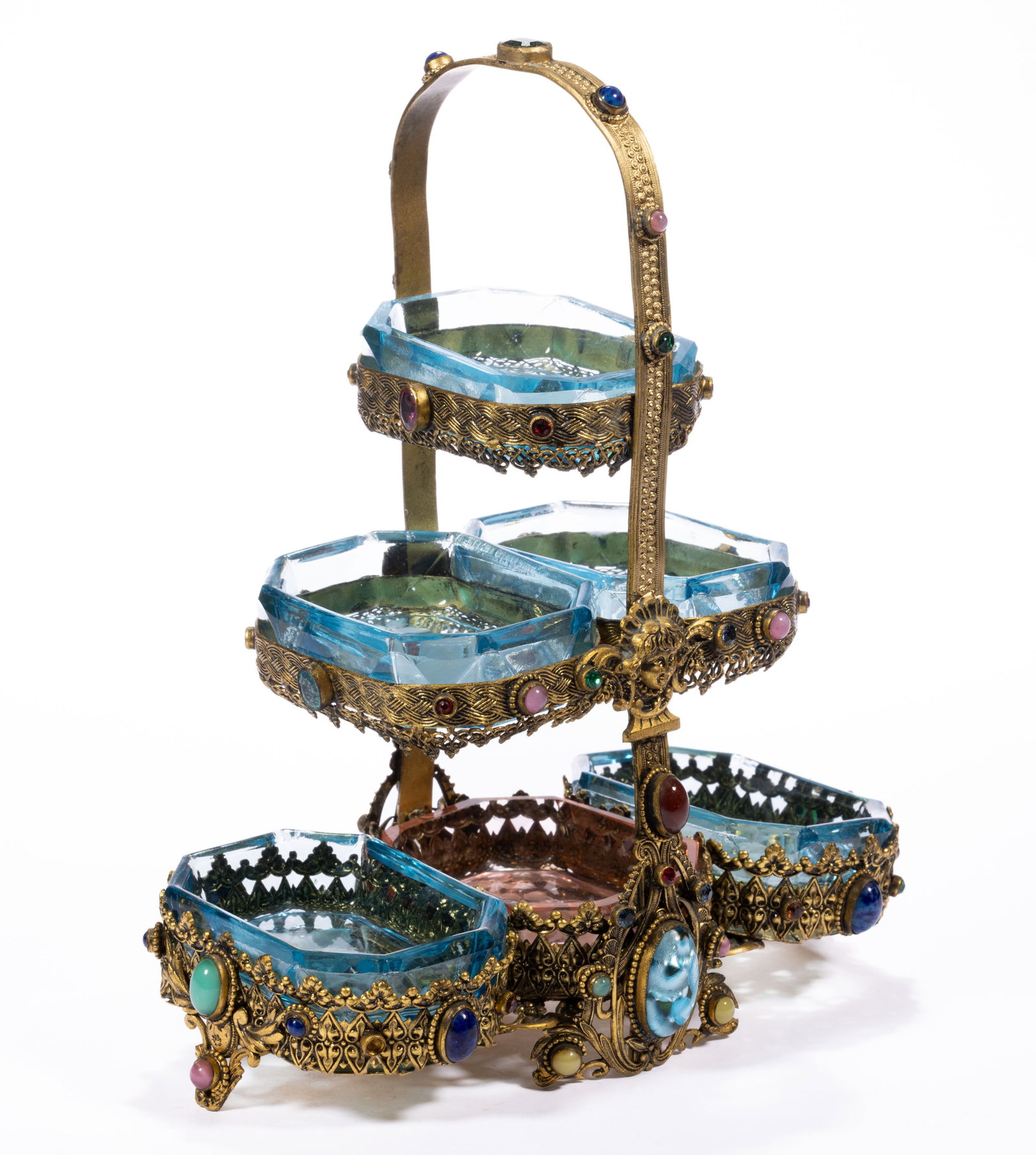 INTAGLIO GLASS SET OF SIX SALTS IN STAND: INTAGLIO GLASS SET OF SIX SALTS IN STAND, pyramid-form brass ornamented stand with many applied jewels to the sides and edges with floral ornamentation to the base, holding five blue and one pink matc