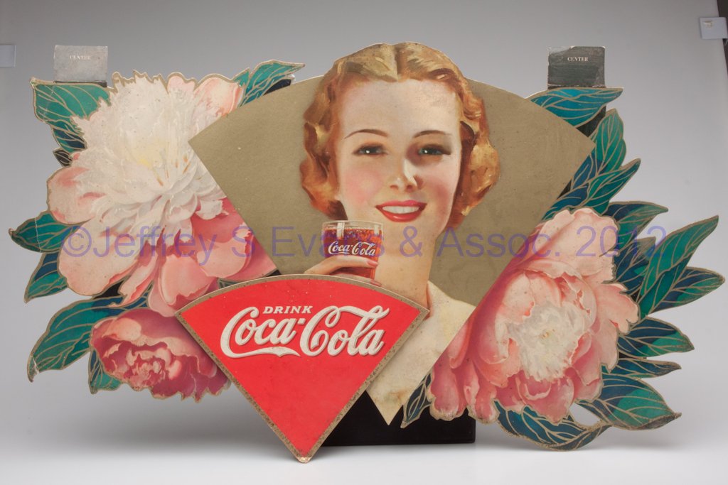 COCA-COLA ADVERTISING SIGN: COCA-COLA ADVERTISING SIGN, lithographed cardboard, depicting a young lady with a glass of Coke surrounded by flowers, upper hanging tabs. Mid 20th century. 20 1/2" x 33". Provenance: From a long-esta