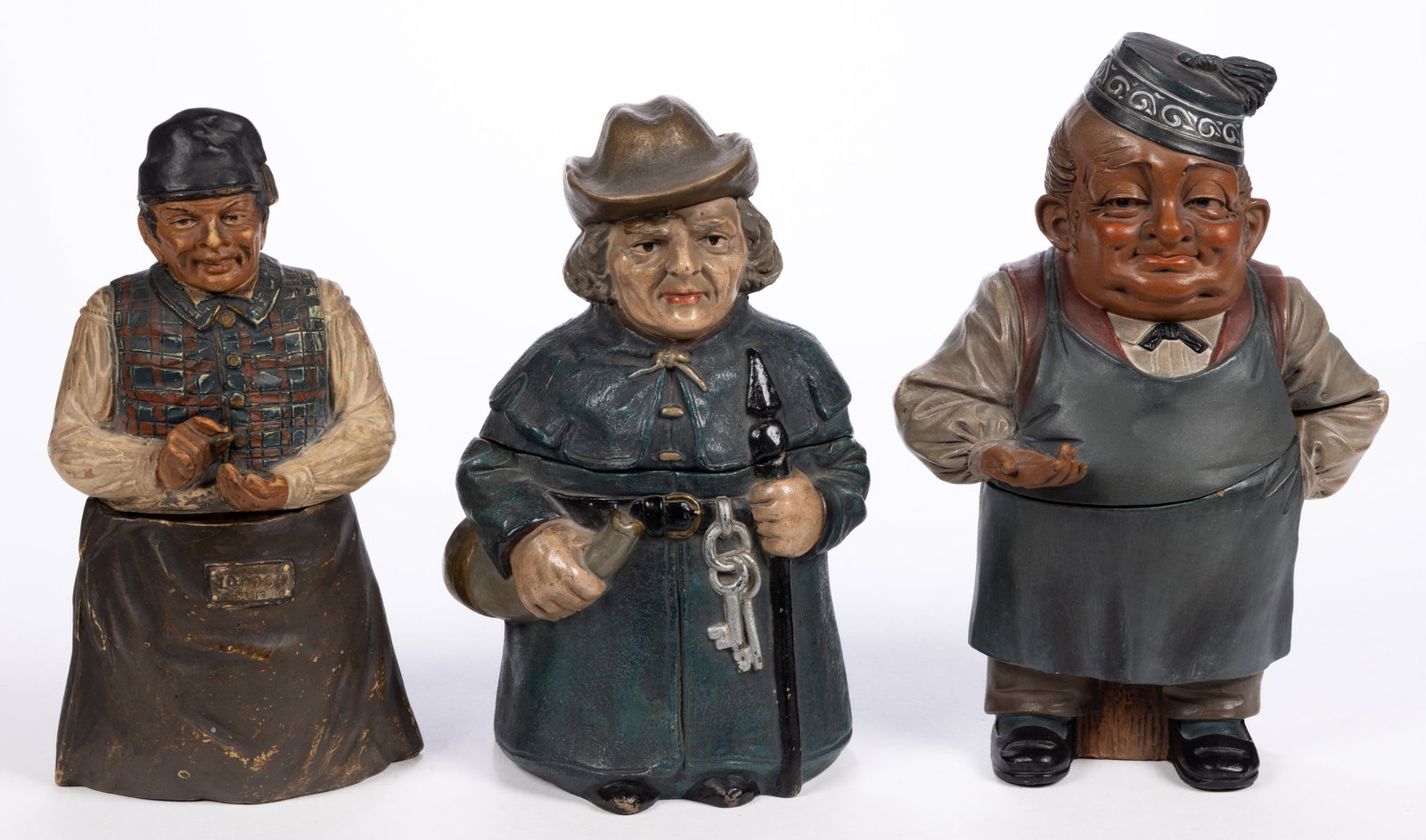 CONTINENTAL CERAMIC OCCUPATIONAL FIGURAL HUMIDORS, LOT OF THREE: CONTINENTAL CERAMIC OCCUPATIONAL FIGURAL HUMIDORS, LOT OF THREE, comprising one example depicting a jailer with a ring of keys to belt and holding a horn and two money collectors including one fe