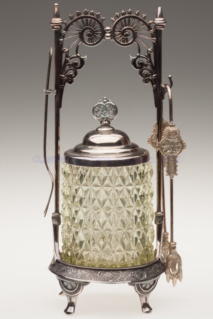 EAPG PRESSED DIAMOND VARIANT PICKLE CASTER: EAPG PRESSED DIAMOND VARIANT PICKLE CASTER, vaseline (uranium), factory polished table ring, Wilcox #94 quadruple-plate stand with fork and tongs. Fourth quarter 19th century. 11" HOA, 4" H jar. Prove