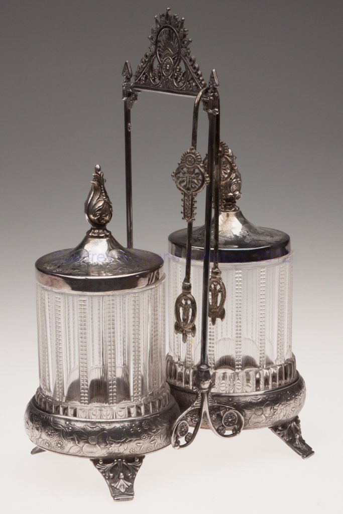 PRESSED GLASS DOUBLE PICKLE CASTER: PRESSED GLASS DOUBLE PICKLE CASTER, colorless, Barbour Bros. quadruple-plate stand with tongs. Fourth quarter 19th century. 11 1/2" HOA, 4 1/4" H jars. Provenance: From the collection of the late Marj