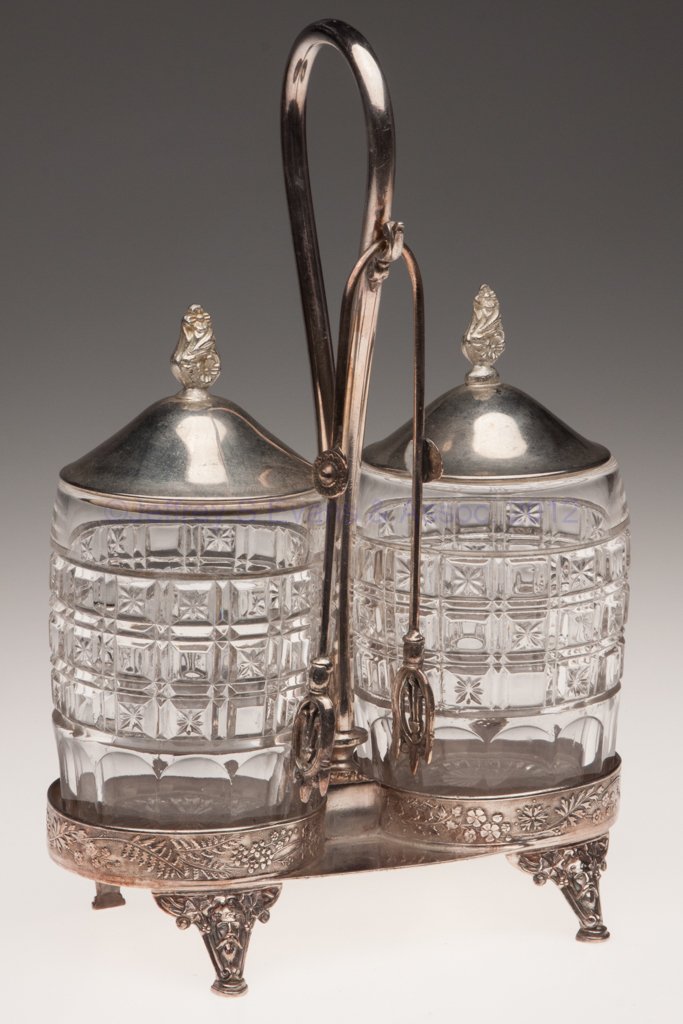 PRESSED GLASS DOUBLE PICKLE CASTER: PRESSED GLASS DOUBLE PICKLE CASTER, colorless, Meriden quadruple-plate stand with tongs. Fourth quarter 19th century. 10" HOA, 4 1/8" H jars. Provenance: From the collection of the late Marjorie Hulse