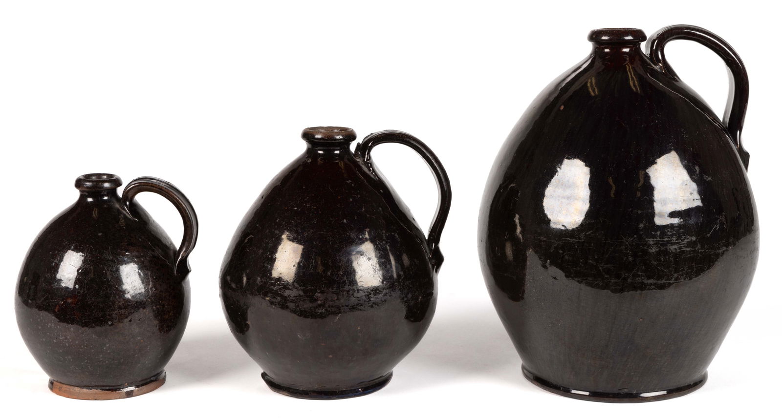 NORTHEASTERN EARTHENWARE / REDWARE JUGS, LOT OF THREE: NORTHEASTERN EARTHENWARE / REDWARE JUGS, LOT OF THREE, graduated, lead- and manganese-glazed, each having exaggerated ovoid form with medial groove strap handle. First half 19th century. 6 3/8&qu