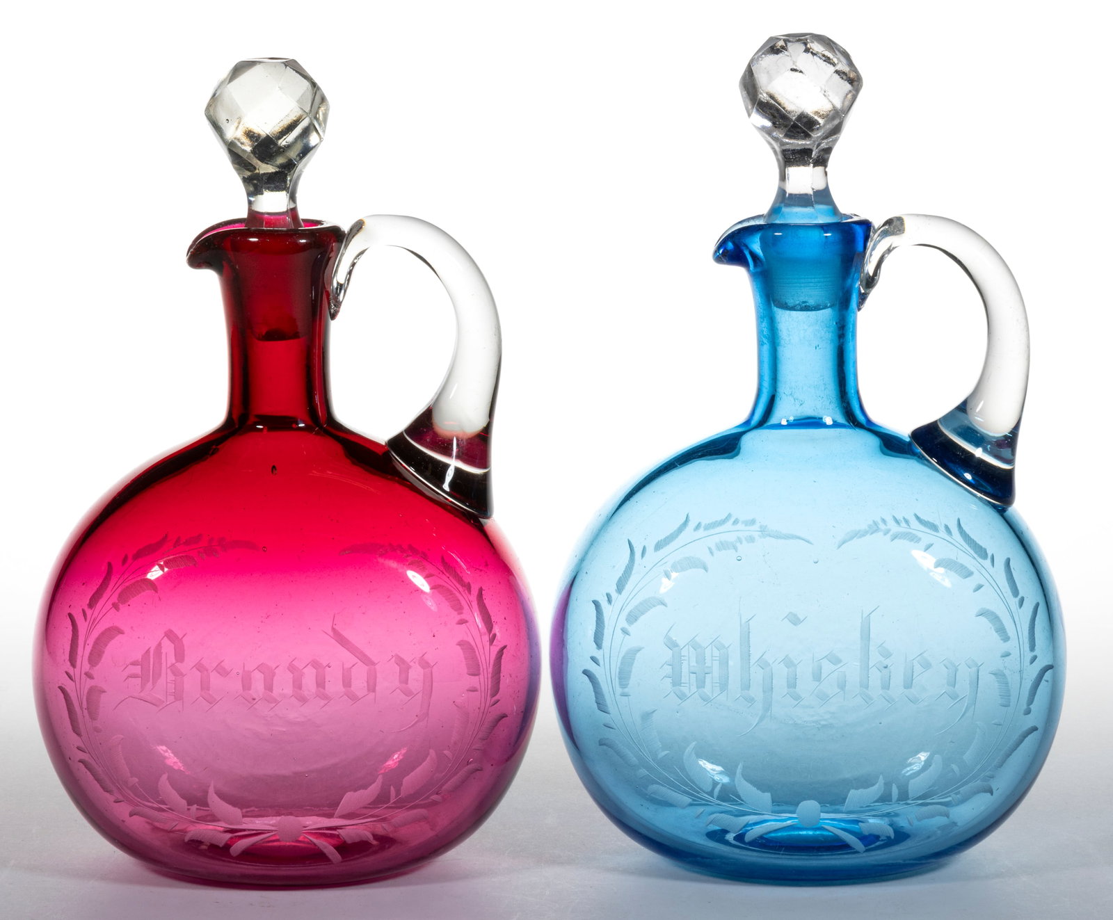 PAIR OF FREE-BLOWN AND ENGRAVED GLASS DECANTERS: PAIR OF FREE-BLOWN AND ENGRAVED GLASS DECANTERS, cased blue and cased cranberry, flat-sided globular bodies with tooled spouts and applied colorless handles, each engraved with a simplistic foliate wr