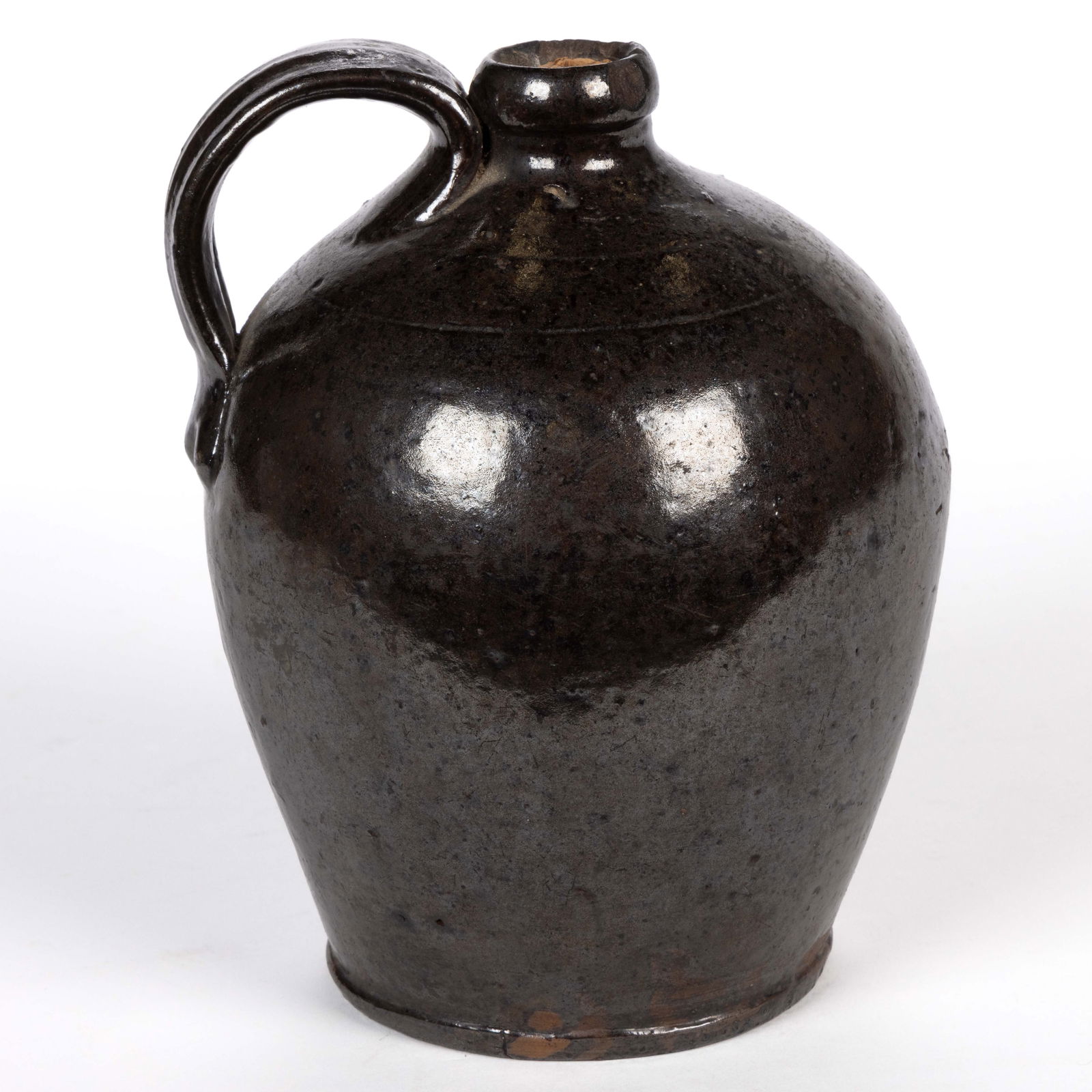AMERICAN EARTHENWARE / REDWARE JUG: AMERICAN EARTHENWARE / REDWARE JUG, lead- and manganese-glazed, ovoid form with two light incised rings to shoulder, applied strap handle with medial groove, and beaded foot. 19th century. 6 1/4"