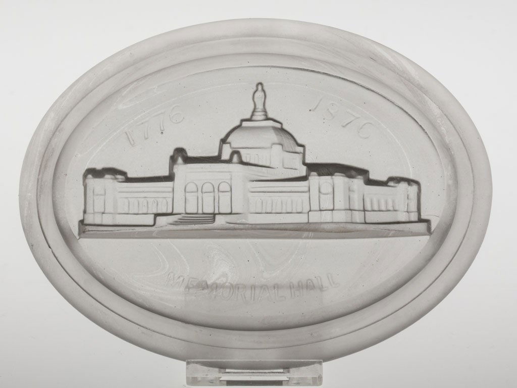 MEMORIAL HALL OVAL PAPERWEIGHT: MEMORIAL HALL OVAL PAPERWEIGHT, frosted except for top surface, pressed intaglio building with dates "1776 1876" above and title below. Probably Gillinder & Sons. Circa 1876. 1 1/8" H, 4" x 5 3/8" bas