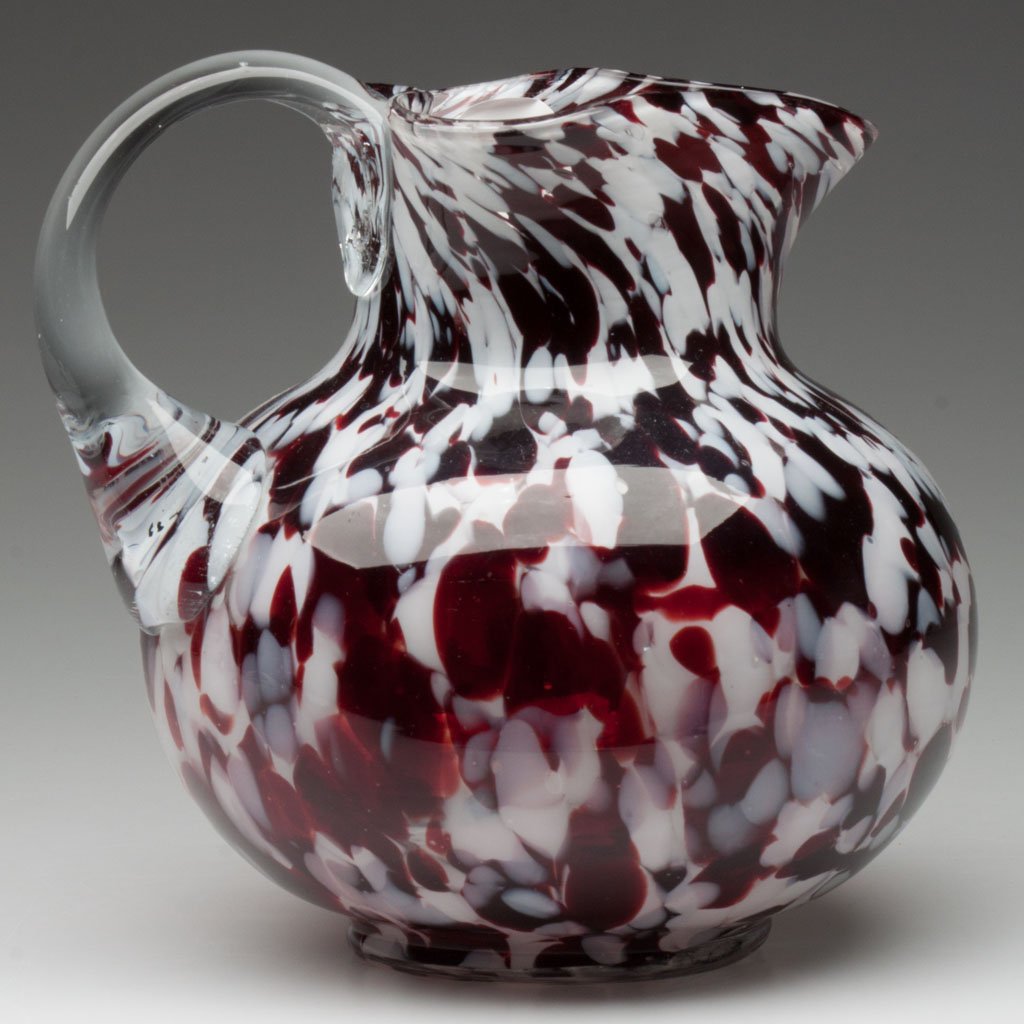 CASED SPATTER WATER PITCHER: CASED SPATTER WATER PITCHER, colorless with maroon and opal flakes, ball form, triangular-form rim, colorless applied handle, polished pontil mark. Late 19th/early 20th century. 6 3/4" H. Provenance: 