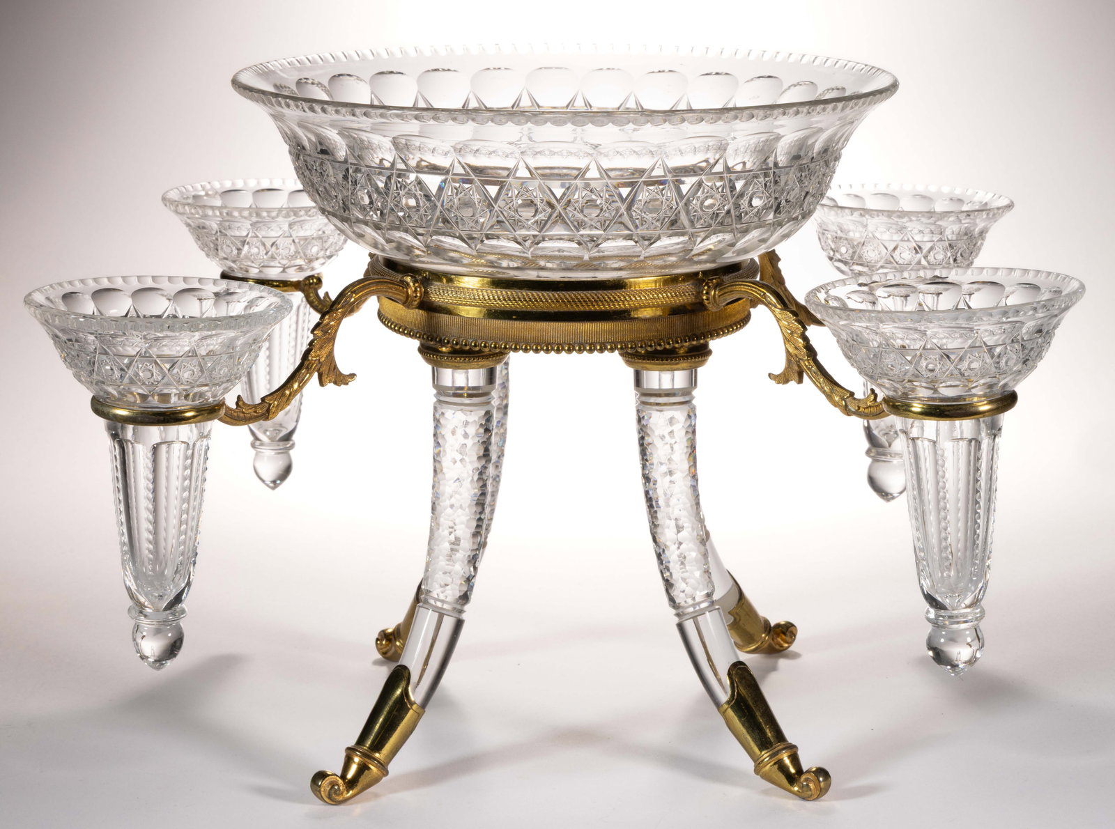 FRENCH GILT-BRASS AND CUT GLASS EPERGNE: FRENCH GILT-BRASS AND CUT GLASS EPERGNE, the circular center bowl fitted in a gilt brass stand with legs cut in the style of rock crystal, four scrolled arms project from the frame and each hold a tru