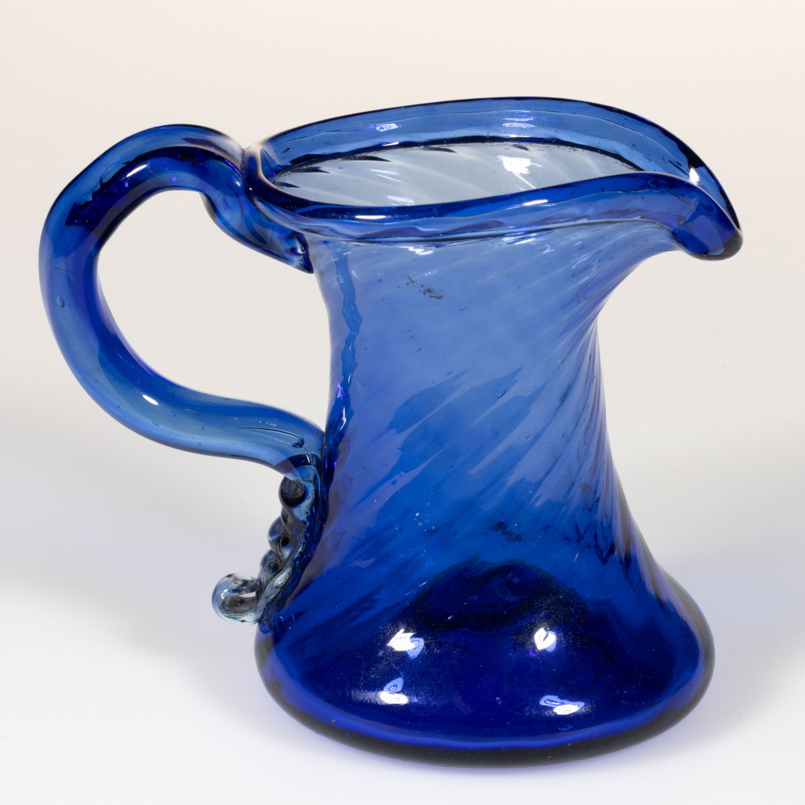 PATTERN-MOLDED GLASS CREAM JUG: PATTERN-MOLDED GLASS CREAM JUG, medium blue, waisted form with 16 ribs swirled to the right, folded over rim, applied handle with two lower crimps and curl, rough pontil mark. Probably English. 19th c