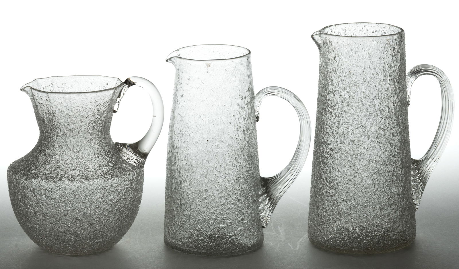 ASSORTED OVERSHOT PITCHERS, LOT OF THREE: ASSORTED OVERSHOT PITCHERS, LOT OF THREE, colorless, comprising an urn-form example with paneled rim and plain applied handle, and two tankard examples, each with an applied reeded handle, rough and p