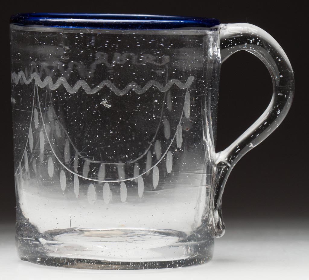 FREE-BLOWN AND ENGRAVED STIEGEL-TYPE MUG: FREE-BLOWN AND ENGRAVED STIEGEL-TYPE MUG, colorless with a gray cast and applied blue rim, cylindrical form with applied handle, swag decoration and faint pontil ring. Late 18th/early 19th century. 3 