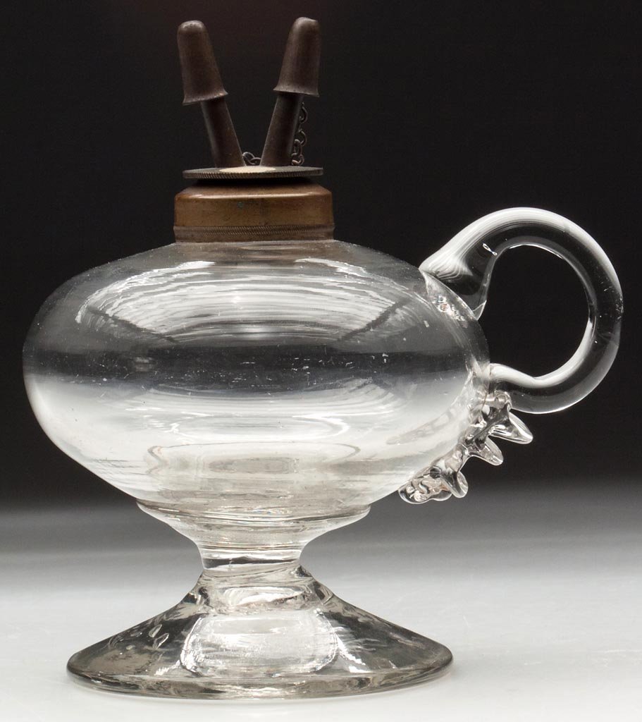 FREE-BLOWN FOOTED HAND LAMP: FREE-BLOWN FOOTED HAND LAMP, colorless, compressed globular font with applied handle featuring a bold, triple-crimp terminal, raised on a flaring, conical foot with rough pontil mark. Brass collar and