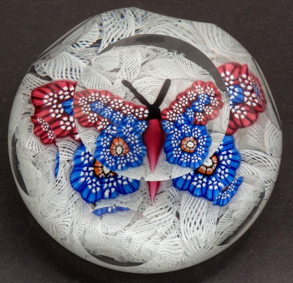 JOHN DEACONS BUTTERFLY FACETTED PAPERWEIGHT: JOHN DEACONS BUTTERFLY FACETTED PAPERWEIGHT, featuring a blue and red millefiori cane-wing butterfly on white muslin ground, polished base with label. Scotland. Second half 20th century. 1 3/4" H, 2 3