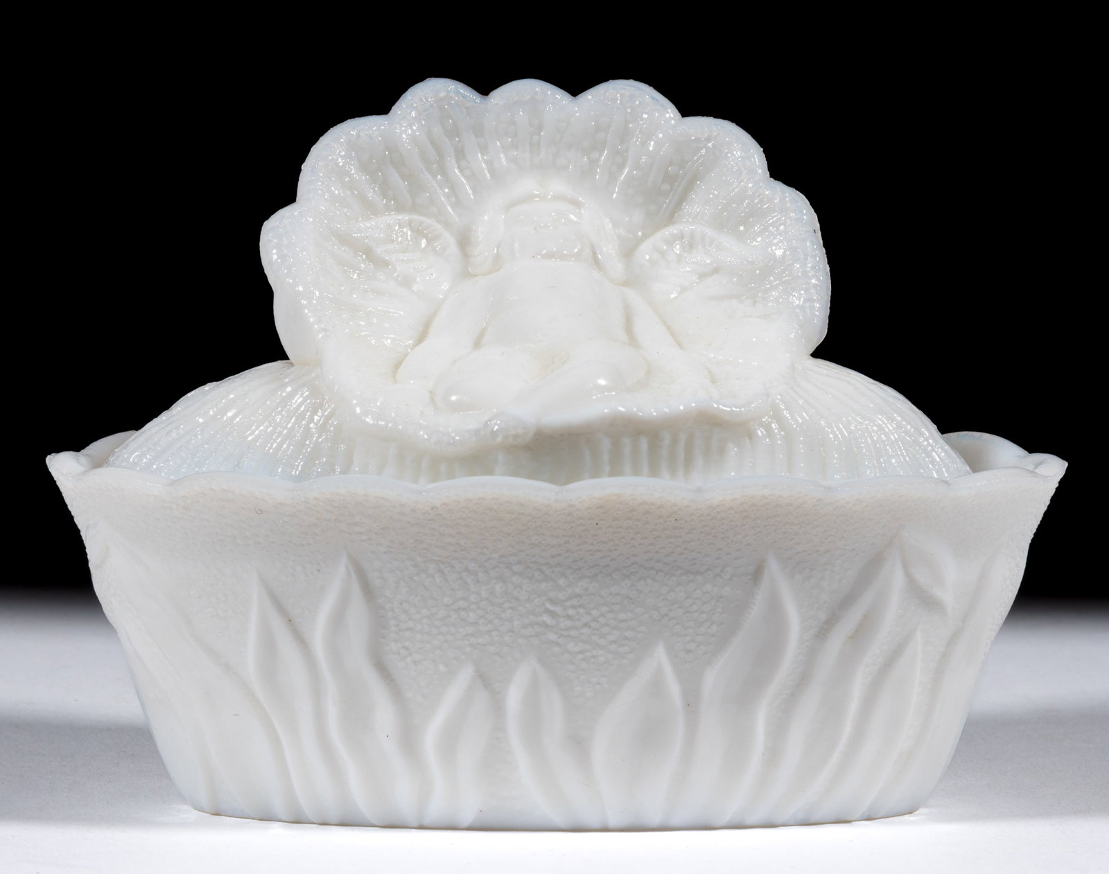 INFANT IN SHELL COVERED DISH: INFANT IN SHELL COVERED DISH, opaque white/milk glass, oval dish with cover featuring a winged infant sitting in a shell, on a scalloped rim rush base. Maker unverified. Late 19th/early 20th cent