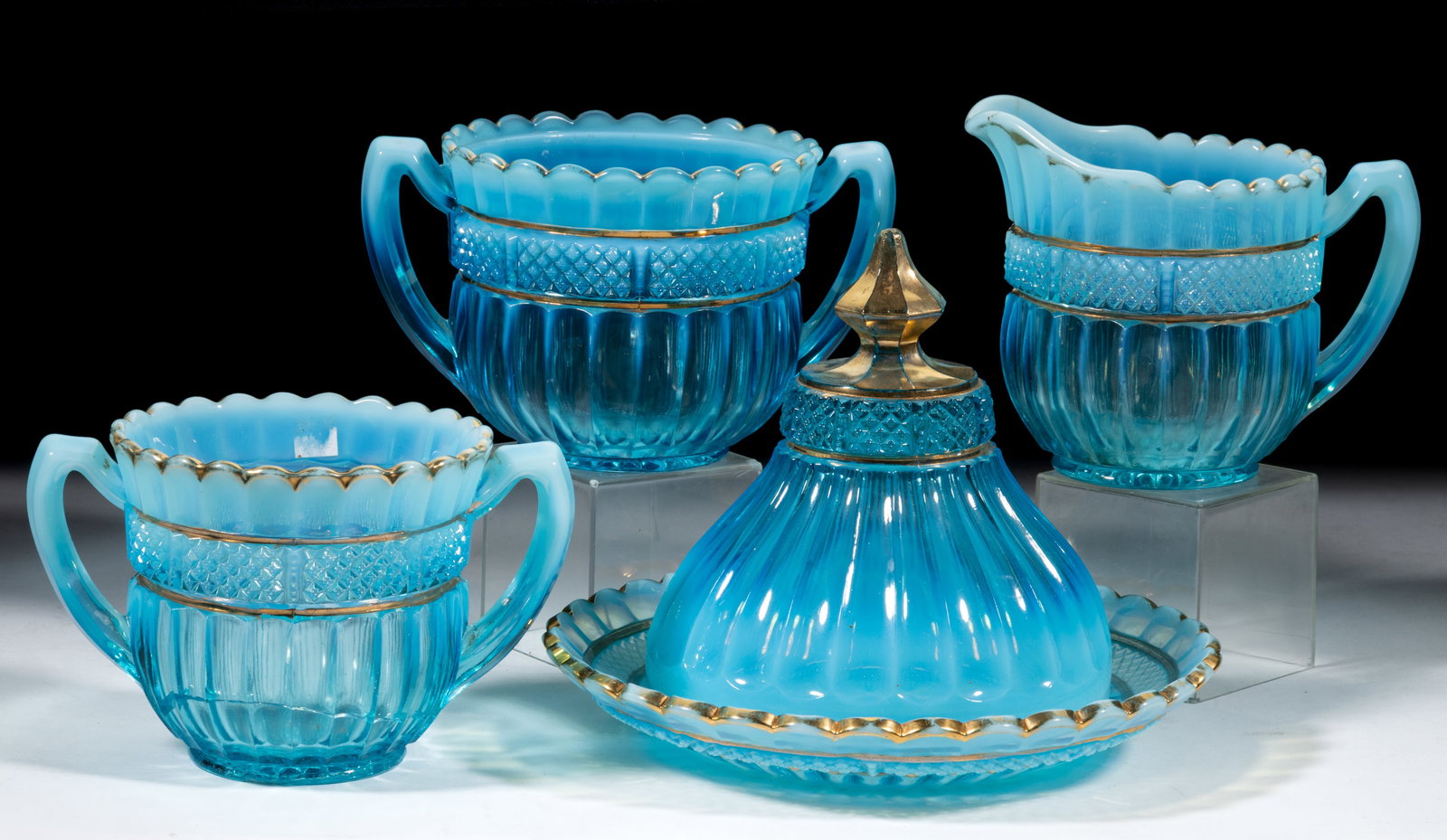 NORTHWOOD LUSTRE FLUTE FOUR-PIECE TABLE SET: NORTHWOOD LUSTRE FLUTE FOUR-PIECE TABLE SET, blue opalescent with gilt decoration, comprising a covered butter dish, an open sugar bowl, a creamer and a spooner, each with the N-in-circle trademark. N