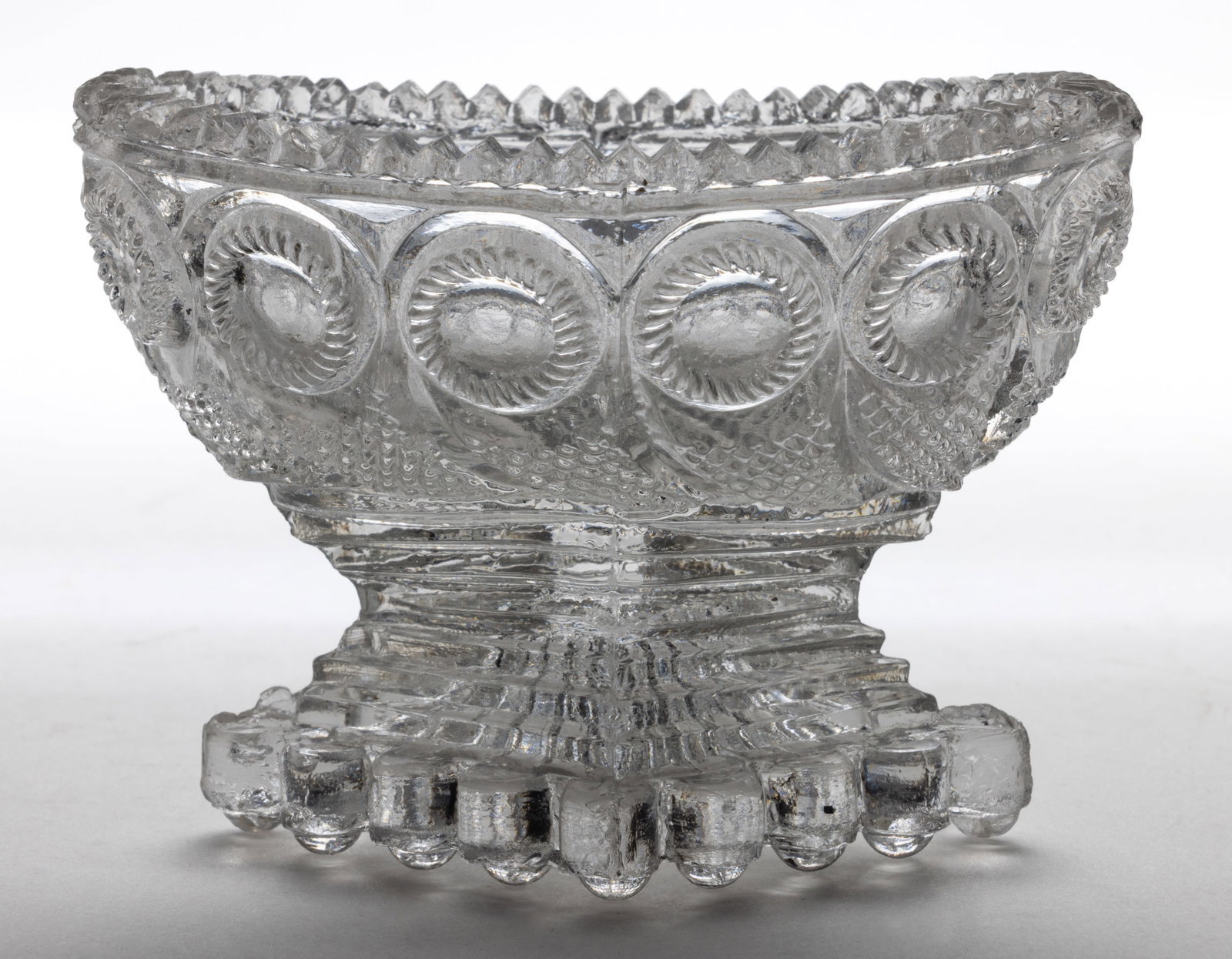 PP-1 PEACOCK EYE PEDESTAL PRESSED OPEN SALT: PP-1 PEACOCK EYE PEDESTAL PRESSED OPEN SALT, colorless, oval boat-like body with a sawtooth rim, raised on a stepped pedestal having a bull's-eye scallop diamond foot with rays to the underside. V