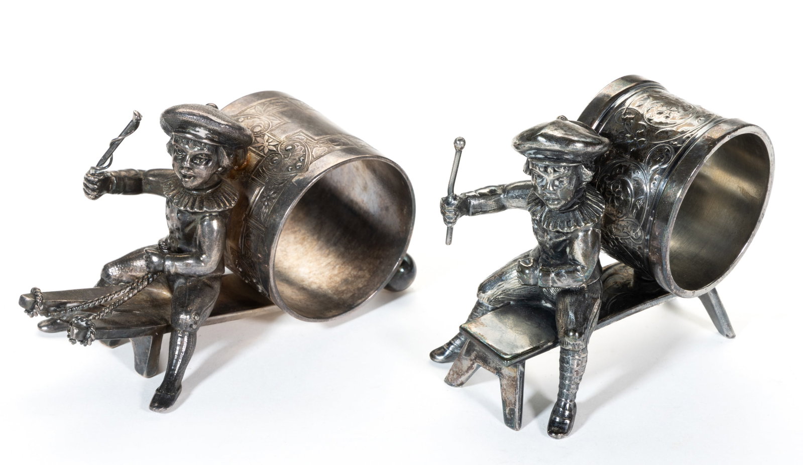 HARTFORD SILVER PLATE CO. AND OTHER KATE GREENAWAY-TYPE BOY FIGURAL SILVER-PLATED NAPKIN RINGS, LOT: HARTFORD SILVER PLATE CO. AND OTHER KATE GREENAWAY-TYPE BOY FIGURAL SILVER-PLATED NAPKIN RINGS, LOT OF TWO, each featuring a boy holding a stick / mallet in one hand sitting on a bench / board, in fro