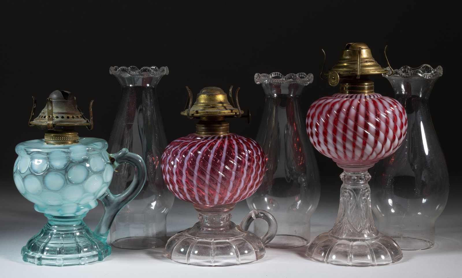 ASSORTED OPALESCENT KEROSENE LAMPS, LOT OF THREE: ASSORTED OPALESCENT KEROSENE LAMPS, LOT OF THREE, comprising of two Sheldon Swirl examples, including a stand example and a footed finger example, each having a cranberry opalescent Reverse Swirl font