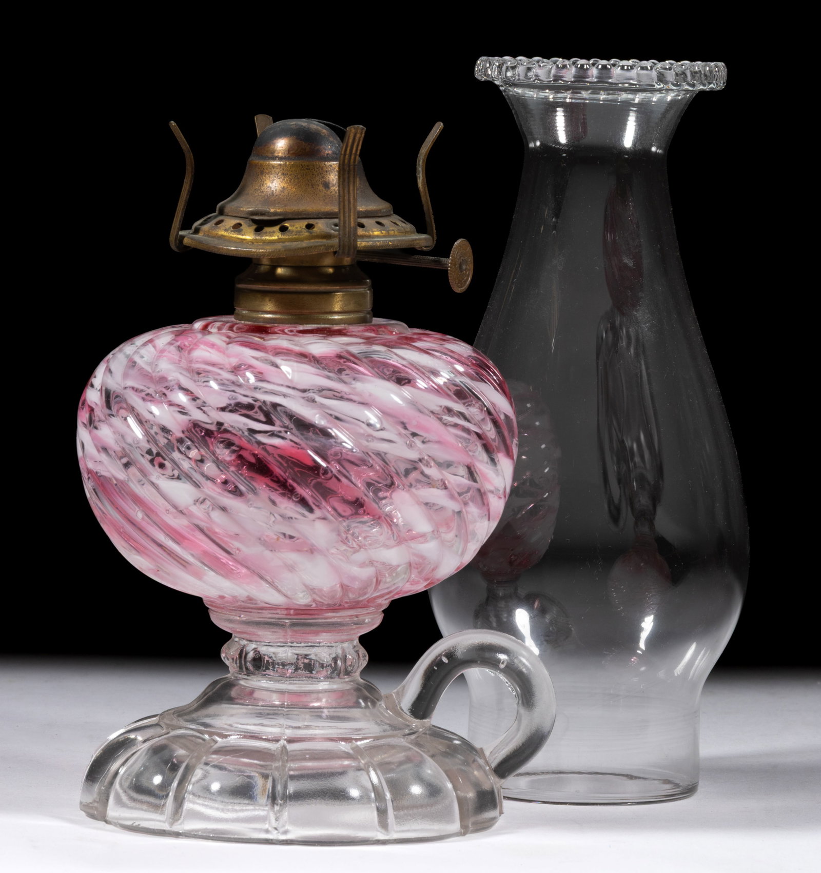 SHELDON SWIRL SPATTER GLASS KEROSENE FOOTED FINGER LAMP: SHELDON SWIRL SPATTER GLASS KEROSENE FOOTED FINGER LAMP, bubbly pink and opal spatter Reverse Swirl font, colorless base, molded handle, No. 1 Taplin-Brown collar. Fitted with a period setup comprisin