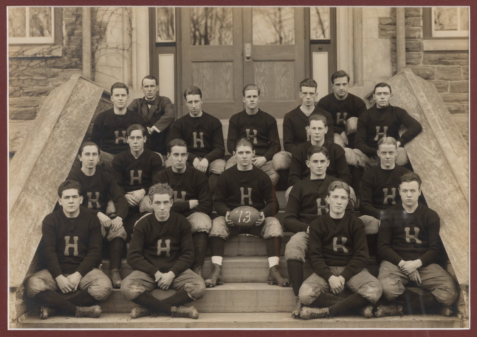 1913 FOOTBALL TEAM GROUP PHOTOGRAPH: 1913 FOOTBALL TEAM GROUP PHOTOGRAPH, gelatin silver print, depicting 16 players, each wearing a V-neck sweater bearing a large "H" to the chest, and one coach, all seated on stone steps in f