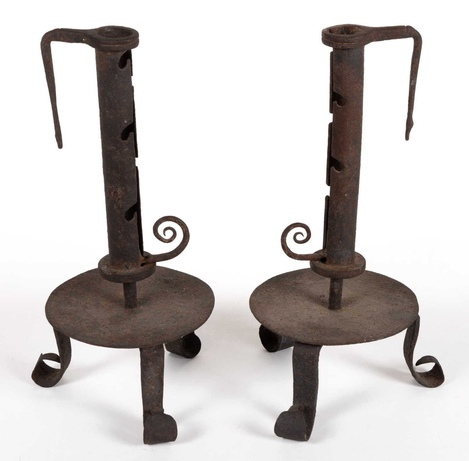 COLONIAL REVIVAL WROUGHT-IRON PAIR OF CANDLESTICKS: COLONIAL REVIVAL WROUGHT-IRON PAIR OF CANDLESTICKS, each with a hanging hook to the adjustable push-up shaft, raised on a circular drip pan and three scrolled feet. 20th century. 11 1/2" H.