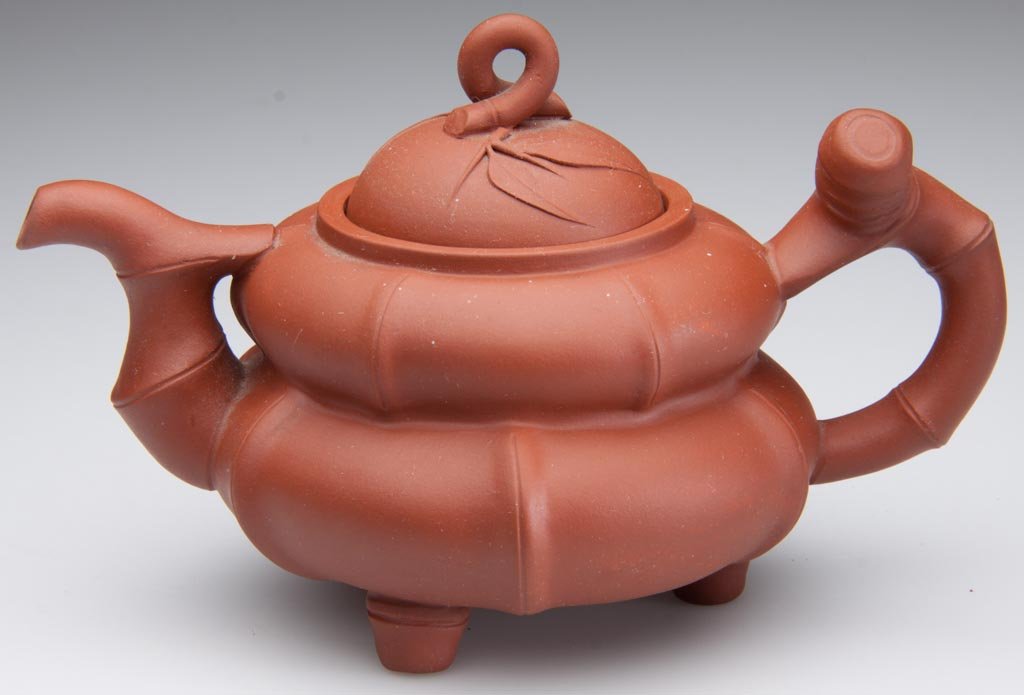 CHINESE YIXING ZISHA CLAY ARTISTIC RED TEAPOT AND COVER: CHINESE YIXING ZISHA CLAY ARTISTIC RED TEAPOT AND COVER, the body formed as a stepped bamboo-form globe on three feet, with bamboo-form handle and spout, the domed cover molded with bamboo leaves, wit