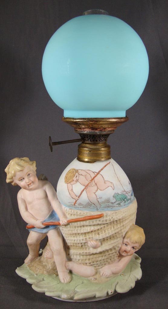 BISQUE DOUBLE CHERUB FIGURAL MINIATURE LAMP: BISQUE DOUBLE CHERUB FIGURAL MINIATURE LAMP, fired décor green, teal, blue, tan, rust and gold trim, with egg shape font and a leaf foot, Blue satin glass ball shade. Period burner. Fourth quarter 19