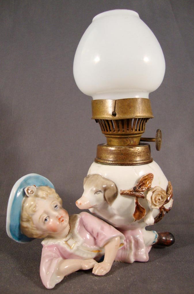 PORCELAIN FIGURAL MINIATURE LAMP: PORCELAIN FIGURAL MINIATURE LAMP, fired décor in blue, pink, brown and gold, girl reclining, egg shape font with pig's head protruding from one side, white Bristol shade. Period burner. Fourth quarte