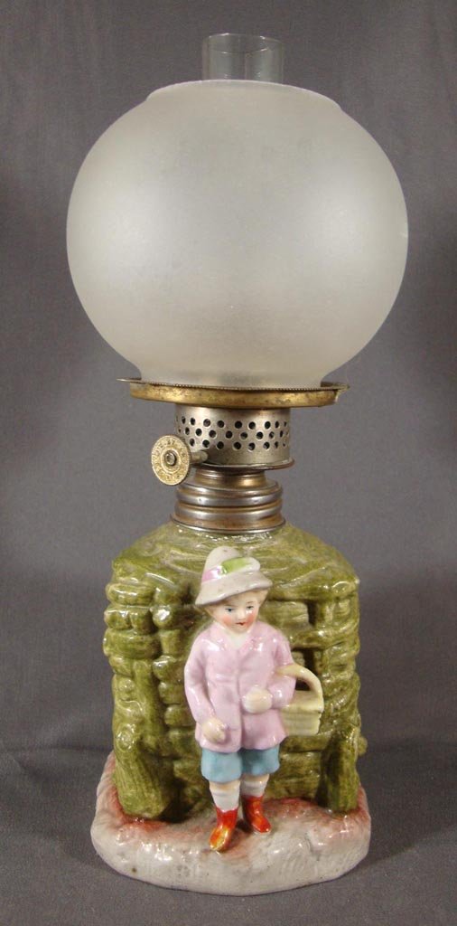 PORCELAIN FIGURAL MINIATURE LAMP: PORCELAIN FIGURAL MINIATURE LAMP, fired décor in green, brown, red, yellow, pink and blue, girl standing before a woven basket, frosted glass ball shade. Period burner. Fourth quarter 19th/early 20th