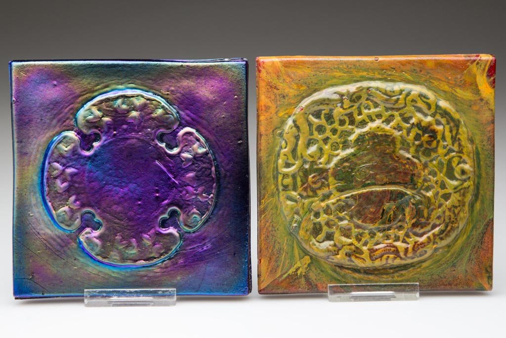 TIFFANY STUDIOS ART GLASS TILES, LOT OF TWO: TIFFANY STUDIOS ART GLASS TILES, LOT OF TWO, comprising an iridescent deep blue example and a mottled green and orange over red example, each molded with design on face and "PAT. APPL'D FOR" and mount