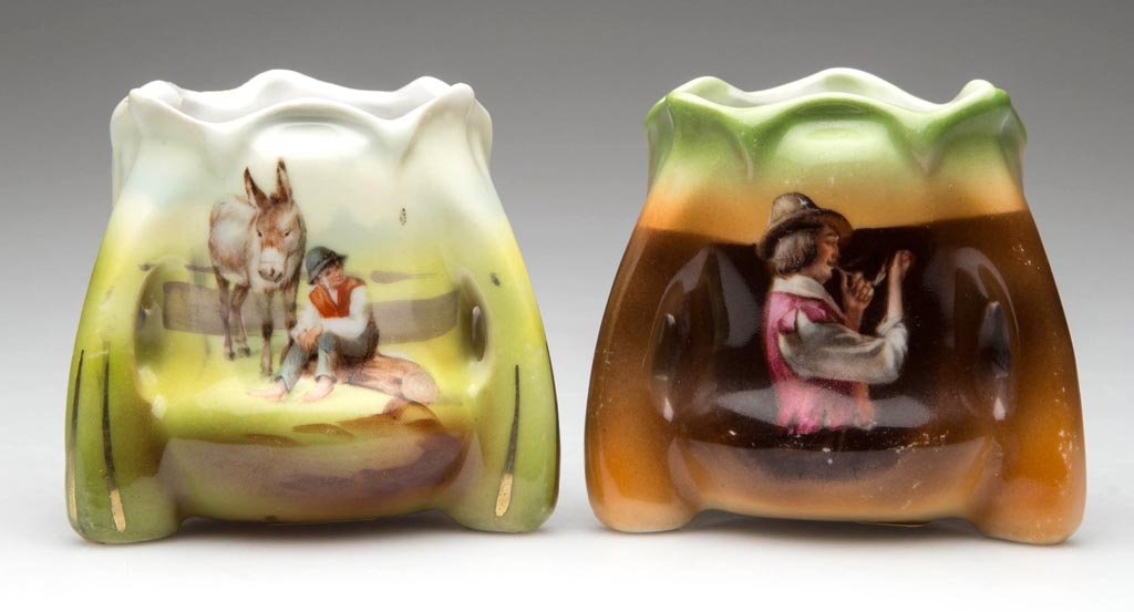 ROYAL BAYREUTH PORCELAIN TOOTHPICK HOLDERS, LOT OF TWO: ROYAL BAYREUTH PORCELAIN TOOTHPICK HOLDERS, LOT OF TWO, each with scalloped rims flaring toward the base and three low handles forming the foot, comprising a "Pipe Smoker" and the second featuring a b