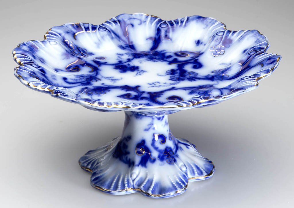 ENGLISH STAFFORDSHIRE POTTERY FLOW BLUE CAKE STAND /: ENGLISH STAFFORDSHIRE POTTERY FLOW BLUE CAKE STAND / TAZZA, the shell-molded dish with scalloped edges on a flaring, hepta-lobe foot, printed with scrolling foliage under a blue border, with gilt edge