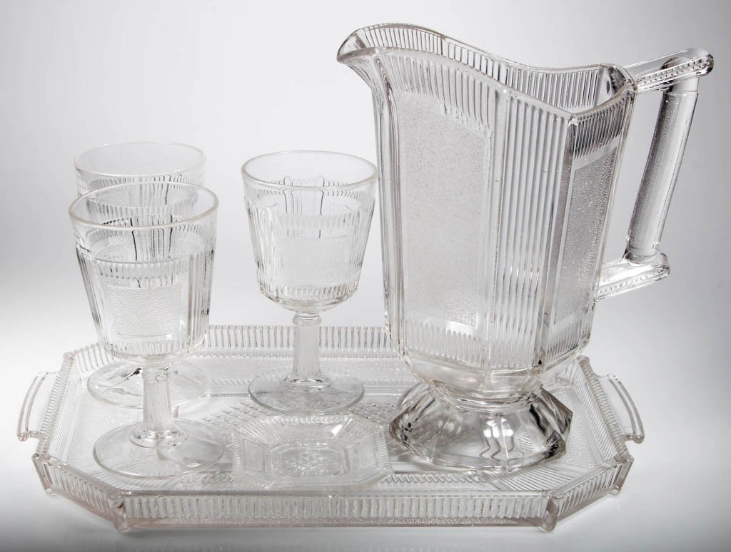 PLEAT AND PANEL TABLE ARTICLES, LOT OF SIX: PLEAT AND PANEL TABLE ARTICLES, LOT OF SIX, colorless, comprising a water pitcher, tray, individual butter pat, and three goblets. Fourth quarter 19th century. Pitcher 9 1/2" H, goblets 6" H, tray 14