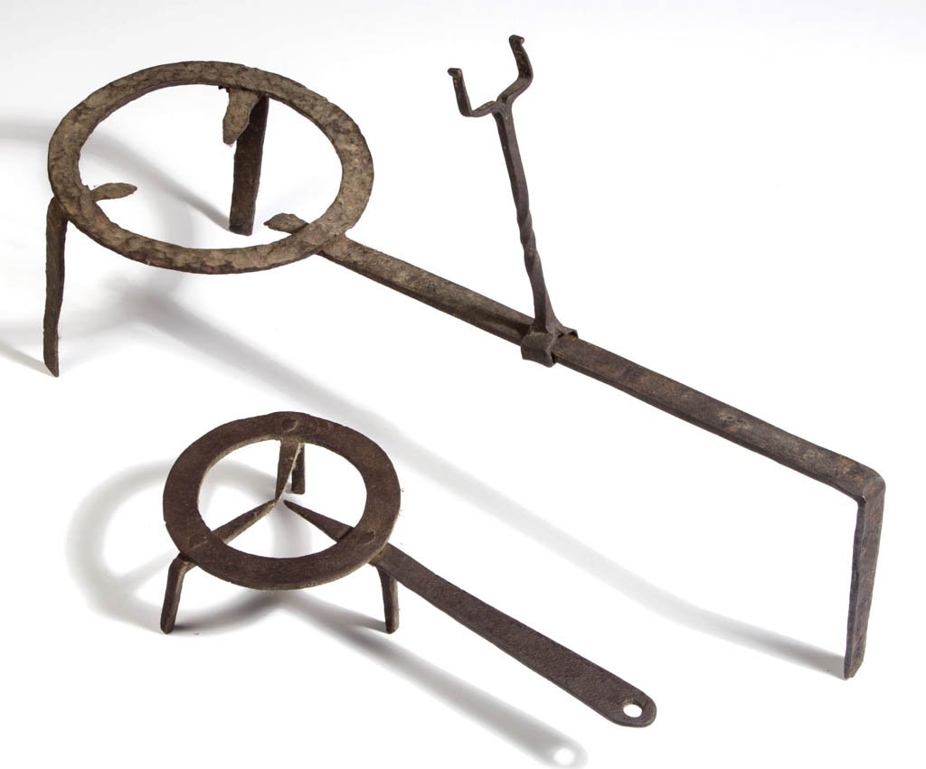 WROUGHT-IRON HEARTH TRIVETS, LOT OF TWO: WROUGHT-IRON HEARTH TRIVETS, LOT OF TWO, comprising an example with adjustable fork-like support, raised on three legs with slightly flared feet; and an example with circular frame enclosing three tap