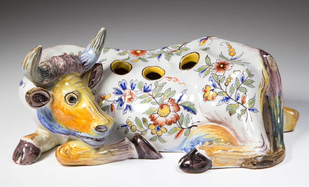CONTINENTAL FAIENCE FIGURAL FLOWER-HOLDER IN THE FORM: CONTINENTAL FAIENCE FIGURAL FLOWER-HOLDER IN THE FORM OF A RECUMBENT COW, with her foreleg bent, head turned to face her udders, each side pierced with three holes, glass eyes. Marked to underside. 20