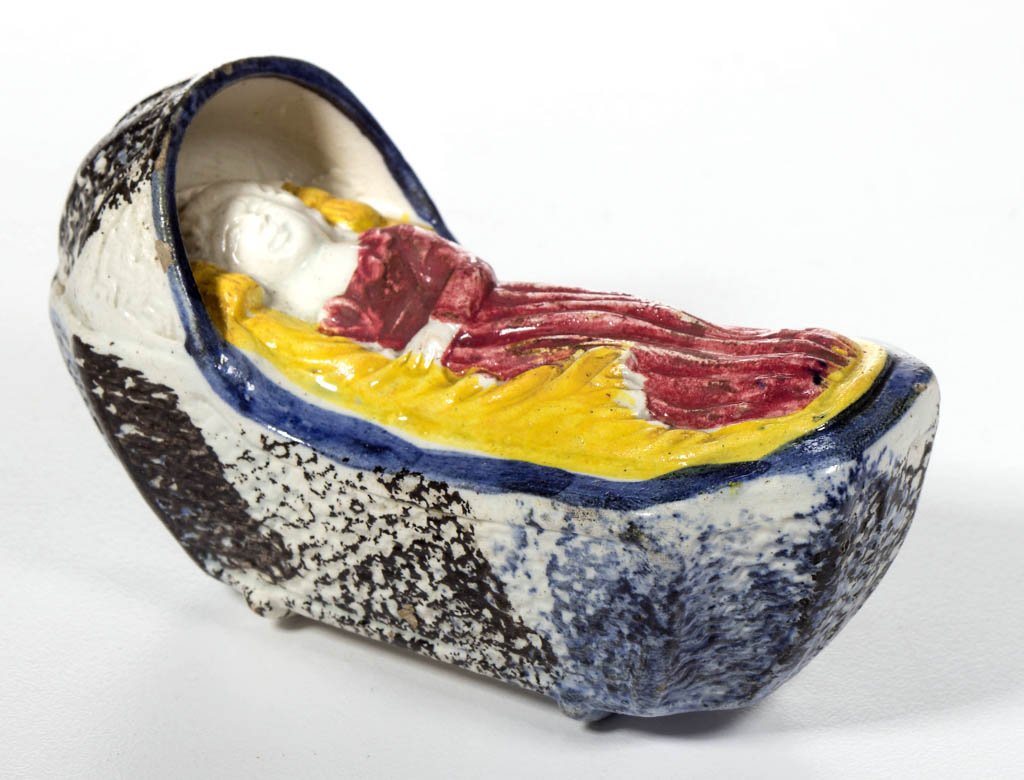 ENGLISH STAFFORDSHIRE POTTERY PEARLWARE PRATTWARE-TYPE: ENGLISH STAFFORDSHIRE POTTERY PEARLWARE PRATTWARE-TYPE BABY IN CRADLE FIGURE, the basketweave-molded hood and bed molded on two rockers, enclosing a small figure of a baby lying asleep, in a red dress