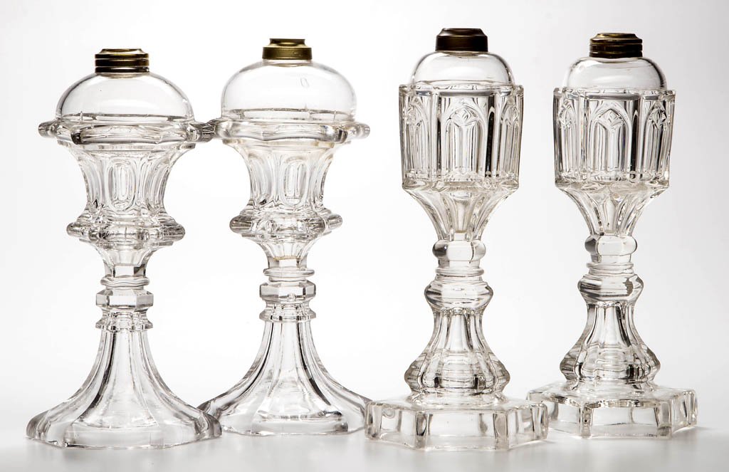 ASSORTED PRESSED PATTERN WHALE OIL / FLUID STAND LAMPS,: ASSORTED PRESSED PATTERN WHALE OIL / FLUID STAND LAMPS, LOT OF FOUR, colorless, comprising near pairs of Waisted Loop and Gothic Arch, each raised on a hexagonal standard and with wafer construction. 