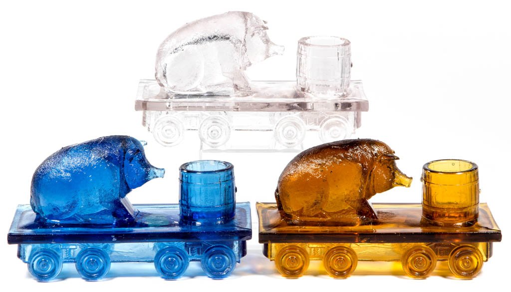 PIG ON A FLAT CAR NOVELTY TOOTHPICK / MATCH HOLDERS,: PIG ON A FLAT CAR NOVELTY TOOTHPICK / MATCH HOLDERS, LOT OF THREE, amber, blue, and colorless. Bellaire Goblet Co. Circa 1880s. 3" HOA, 5 1/2" x 2 1/2".Literature: Sanford - Victorian Glass Novelties,