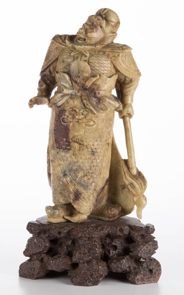 CHINESE SOAPSTONE CARVING OF A FIERCE GUARDIAN /: CHINESE SOAPSTONE CARVING OF A FIERCE GUARDIAN / WARRIOR, the figure standing, holding a fire standard in one hand, his head turned to one side, with a mask carved to the garment at his groin. Fixed t
