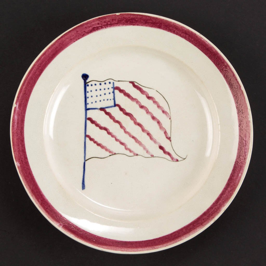 PATRIOTIC "AMERICAN FLAG" DECORATED WHITEWARE CERAMIC: PATRIOTIC "AMERICAN FLAG" DECORATED WHITEWARE CERAMIC PLATE, red, white, and blue, the center hand-painted with a 28-star flag with six diagonal stripes flying from a flag pole, wide-stripe border sur
