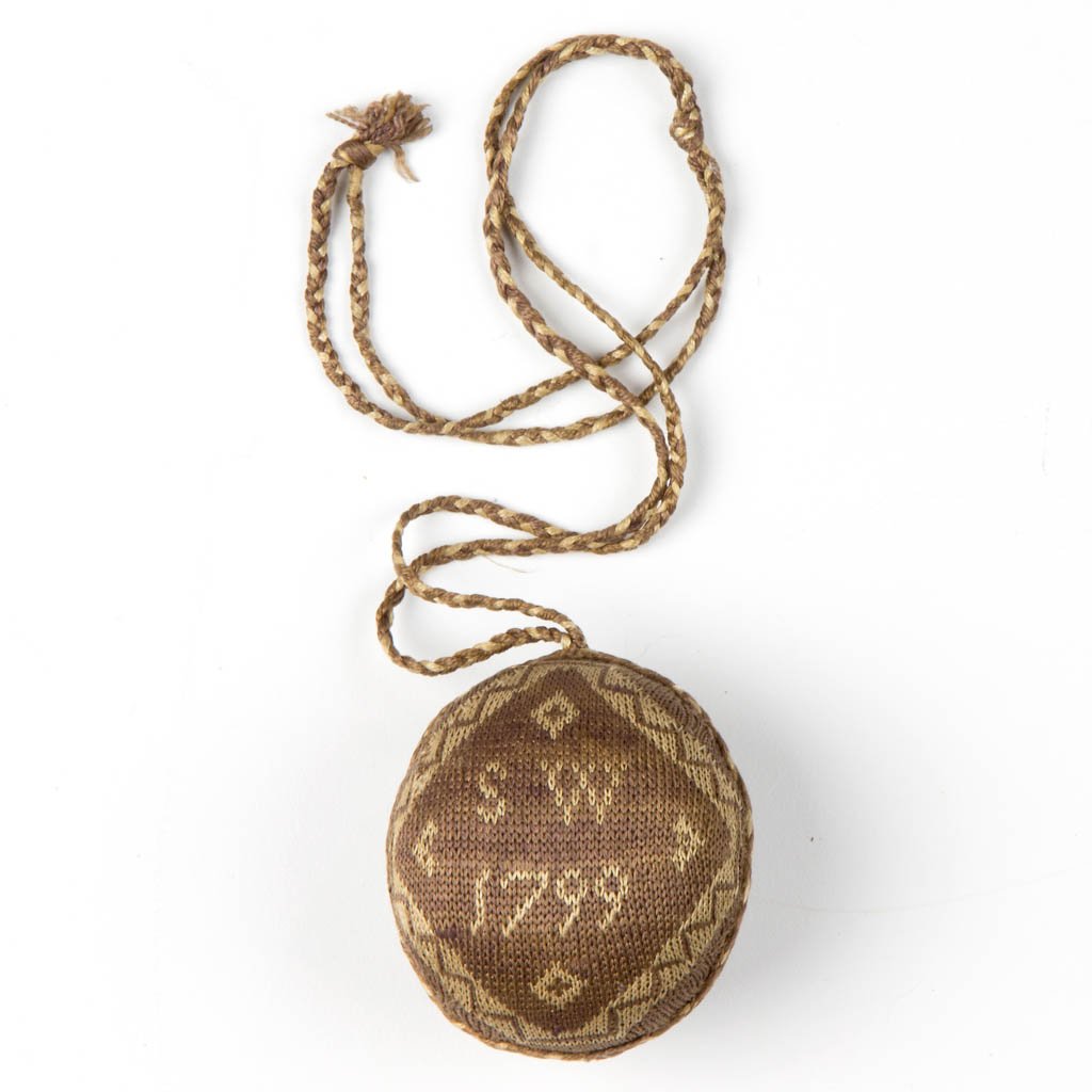 KNITTED INITIALED AND DATED 1799 PIN-BALL CUSHION: KNITTED INITIALED AND DATED 1799 PIN-BALL CUSHION, brown and ecru, braided cord attached and securing two joined halves, one side with "S W / 1799" and reverse with a checkered diamond and tulip-type 