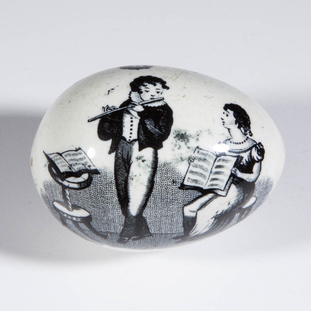 ENGLISH STAFFORDSHIRE TRANSFERWARE CHILD'S EGG: ENGLISH STAFFORDSHIRE TRANSFERWARE CHILD'S EGG, printed in black, depicting a boy taking flute lessons, and a boy with his dog and a basket of birds, lettered "A PRESENT FOR A / GOOD BOY" between scen