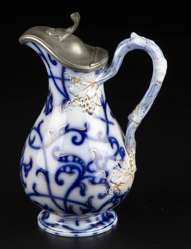ENGLISH IRONSTONE FLOW BLUE SYRUP PITCHER: ENGLISH IRONSTONE FLOW BLUE SYRUP PITCHER, transfer printed, the elongated globular body on foot, molded with grape leaves and fruit at the vine-form handle terminates, transfer-printed in cobalt blue