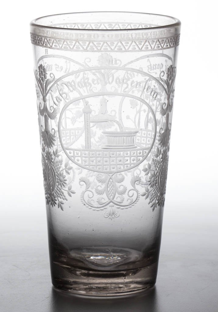 CONTINENTAL FREE-BLOWN AND ENGRAVED TALL BEAKER: CONTINENTAL FREE-BLOWN AND ENGRAVED TALL BEAKER, colorless non-lead glass, tapered form with three numbered reserves featuring hops, grapes, and a water pump, each with inscription above, thick base w