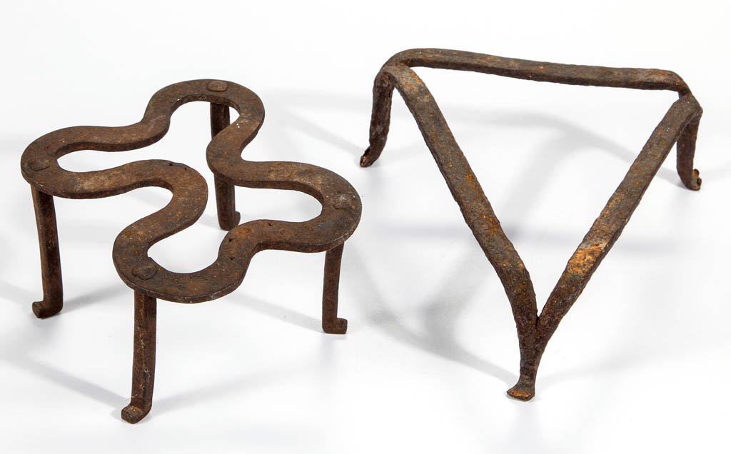 AMERICAN WROUGHT-IRON HEARTH TRIVETS, LOT OF TWO: AMERICAN WROUGHT-IRON HEARTH TRIVETS, LOT OF TWO, comprising a triangular example with three drawn legs and a curvilinear example with four applied legs. Original surfaces. Found in the Shenandoah Val