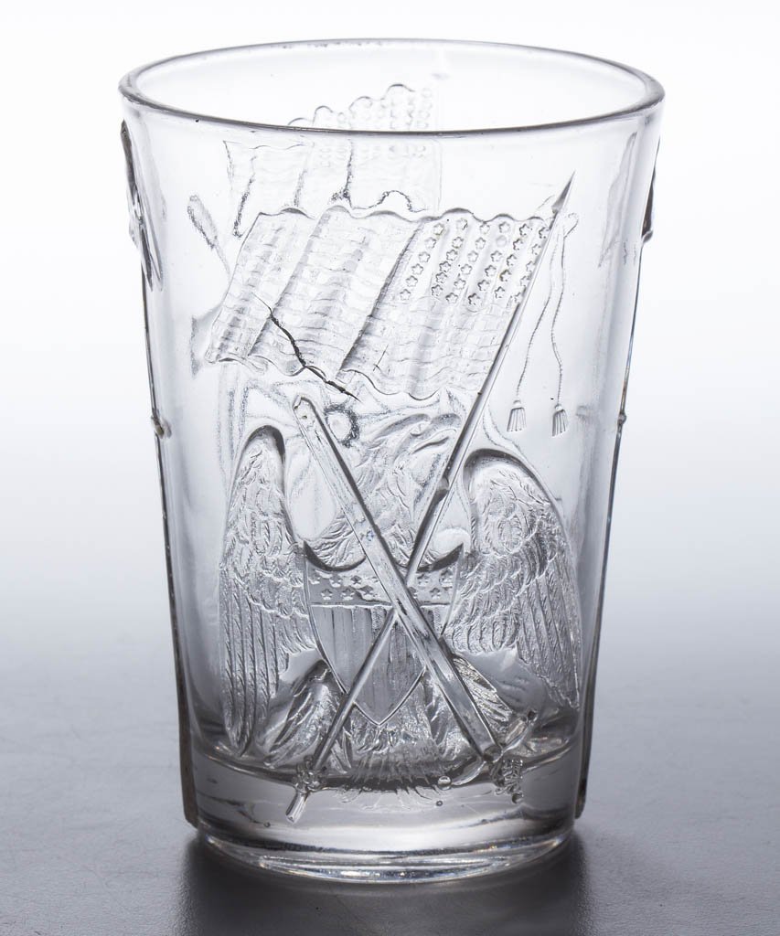 PRESSED CIVIL WAR TUMBLER: PRESSED CIVIL WAR TUMBLER, colorless lead glass, one side embossed with cannon, trench mortar, tamping rod, and cannon balls with flag flying above, reverse featuring a spread-wing American eagle with